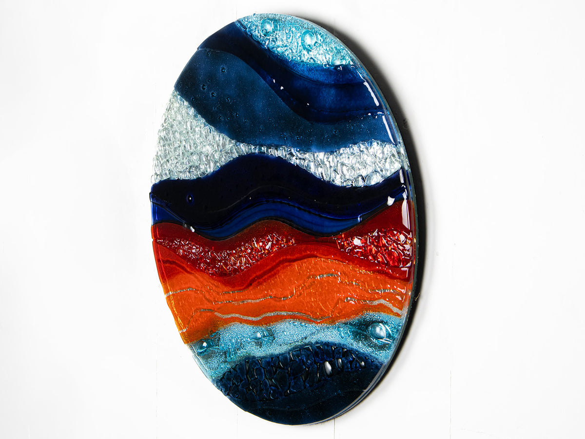 A circular fused glass wall panel featuring abstract wave design in ocean blues and bright oranges, with textured detailing. 