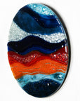 A circular fused glass wall panel featuring abstract wave design in ocean blues and bright oranges, with textured detailing. 