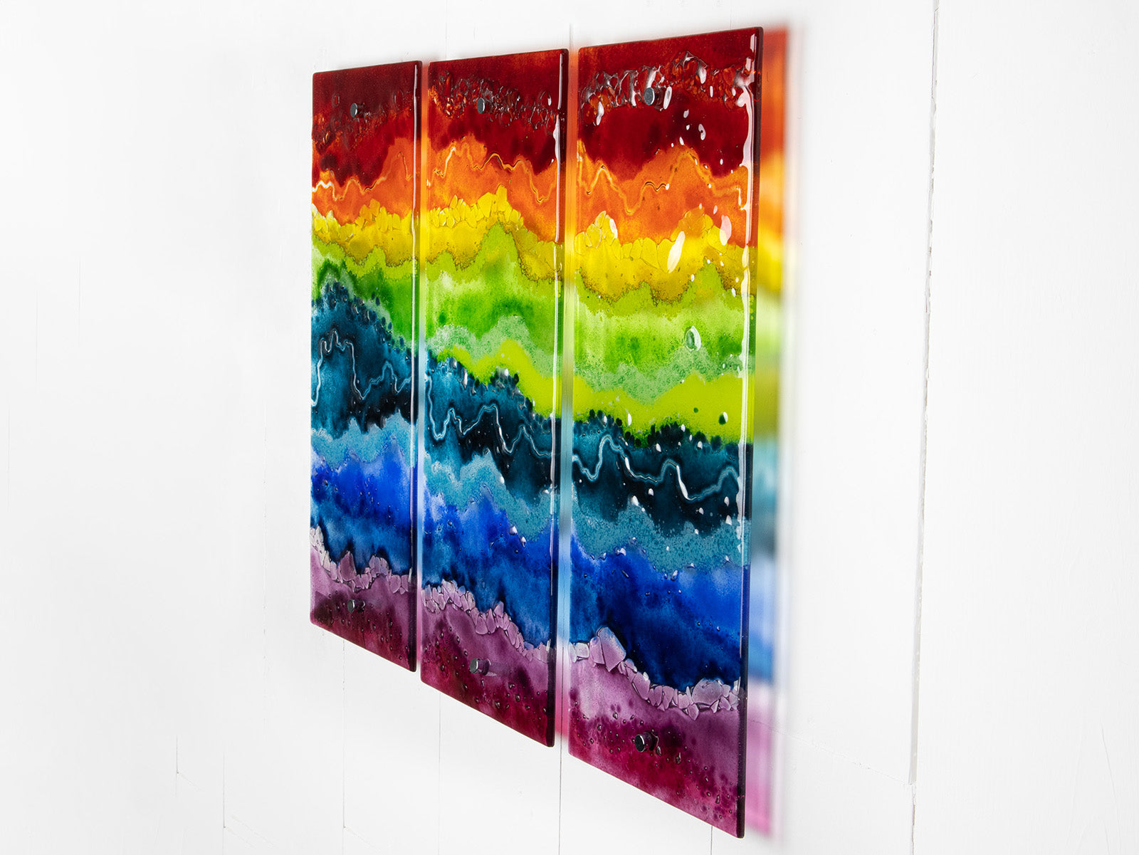 A three-panel vertical fused glass wall art piece featuring wavy horizontal rainbow stripes in red, orange, yellow, green, blue, and purple, mounted on a white wall.