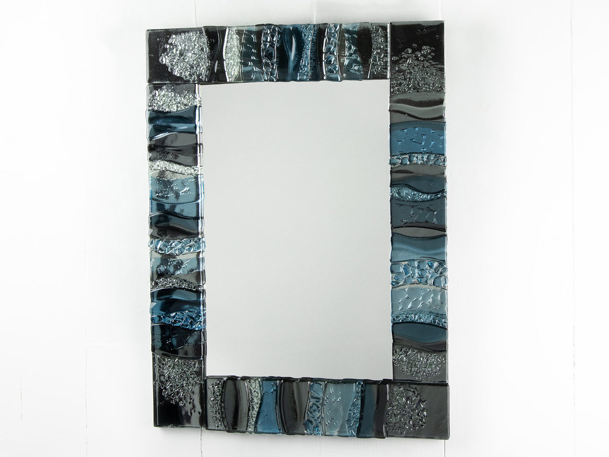 A fused glass oblong mirror, handmade using soft coastal blue tinted and grey glass with delicate texture and a wavy design.