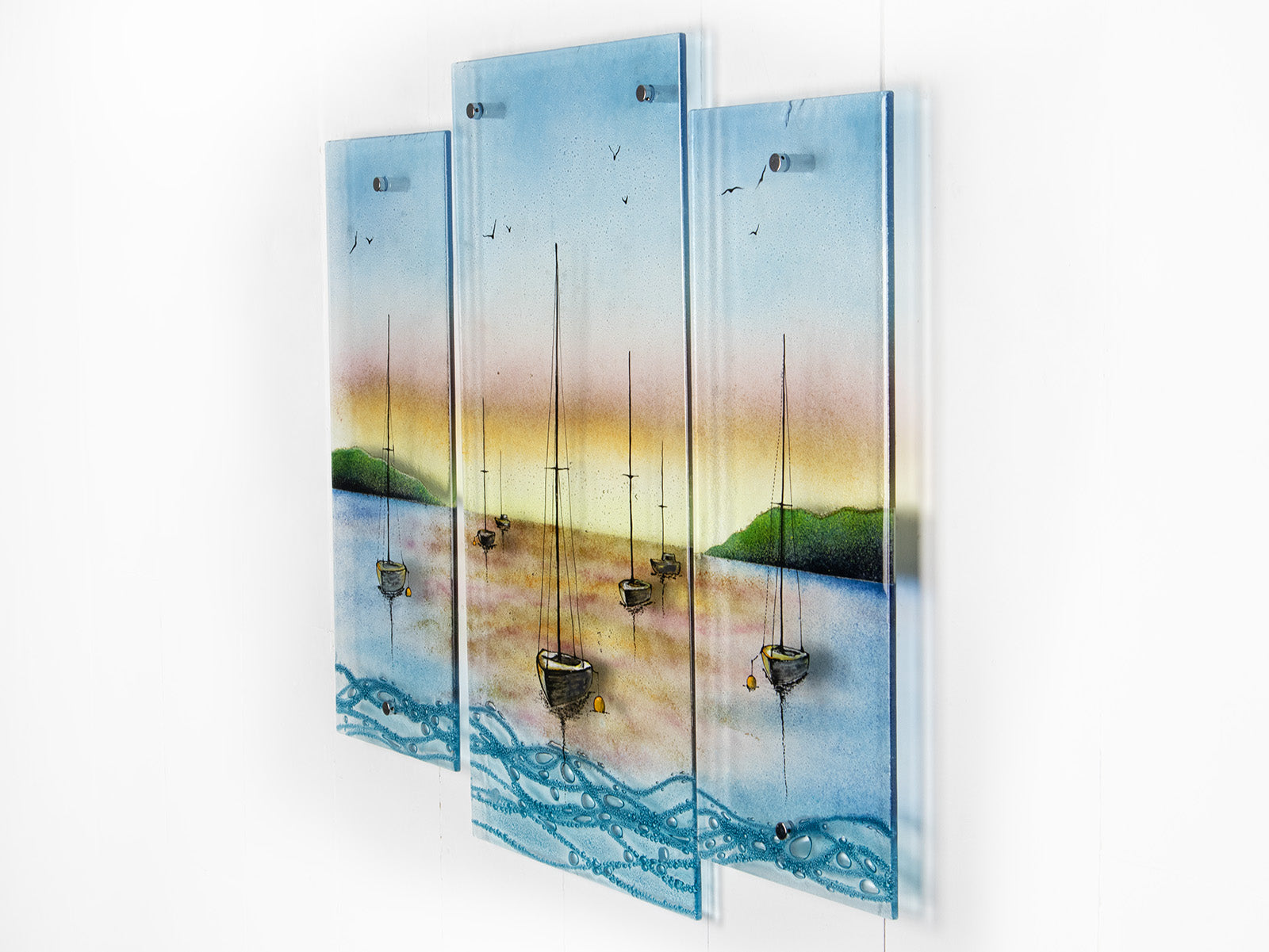 A side image of a fused glass triptych depicting a serene harbour scene with hand-painted sailboats on blue water under a pastel sky with hills in the distance.