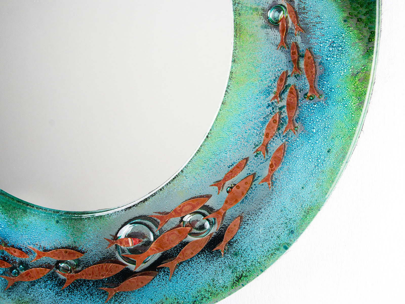 A close up of a circular fused glass round mirror, handmade in vibrant aquas and sea greens, with darting copper fish. 
