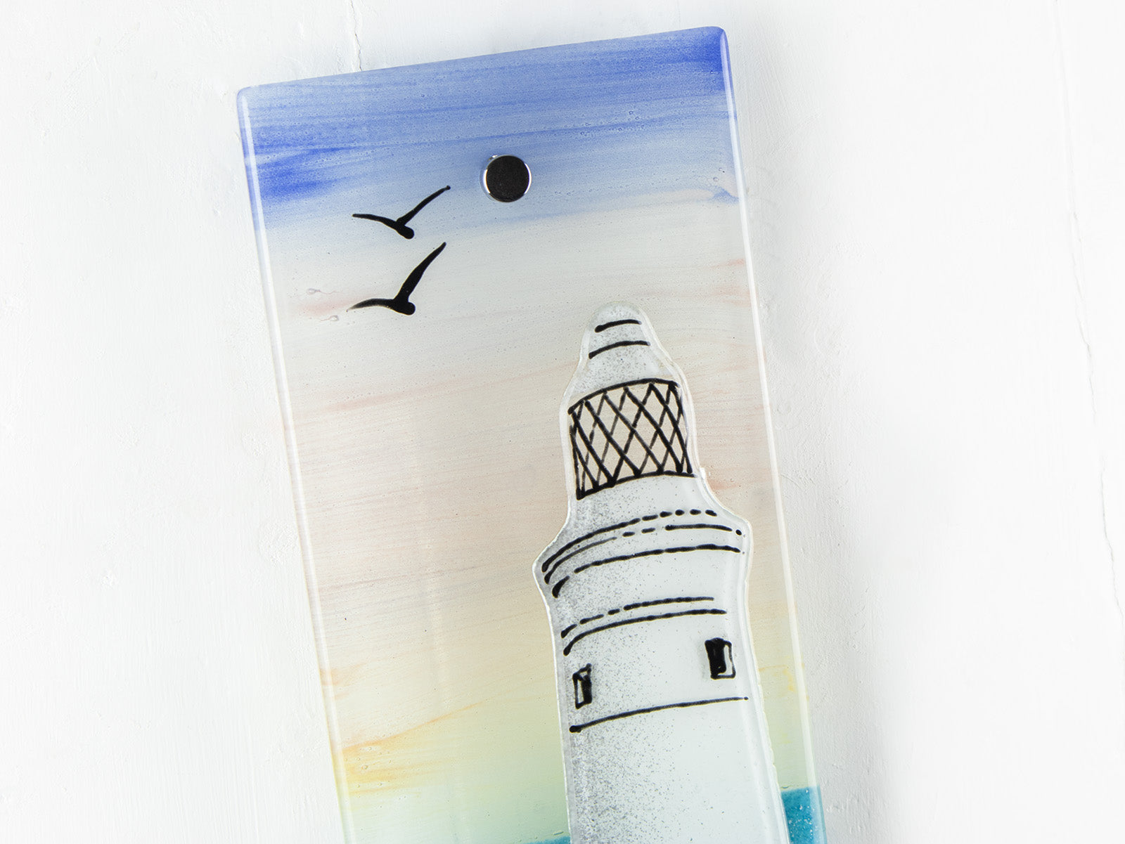 A close up of a fused glass extra small wall panel, featuring a lighthouse design with a very soft sunset pink sky, with soaring birds. A soft coastal piece of wall art. 
