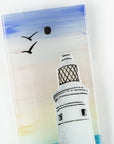 A close up of a fused glass extra small wall panel, featuring a lighthouse design with a very soft sunset pink sky, with soaring birds. A soft coastal piece of wall art. 