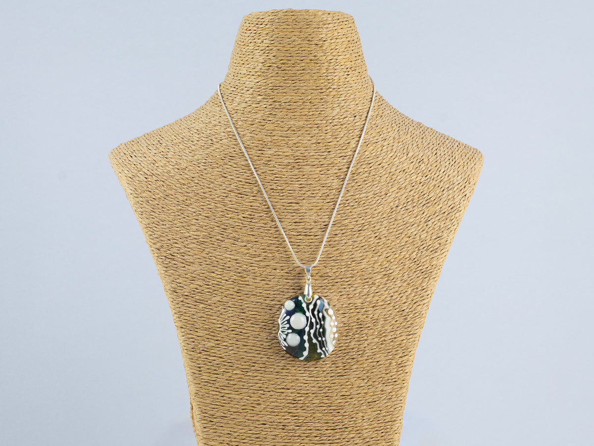 A fused glass pendant necklace, handmade in soft coastal and organic hues, with hand-painted botanical patterns, hung on sterling silver chain. 