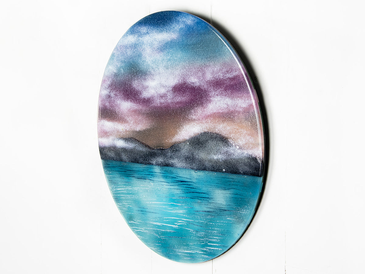 A circular wall art fused glass panel featuring a scenic landscape with a blue and purple sky, misty mountains, and turquoise water. 