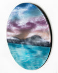 A circular wall art fused glass panel featuring a scenic landscape with a blue and purple sky, misty mountains, and turquoise water. 