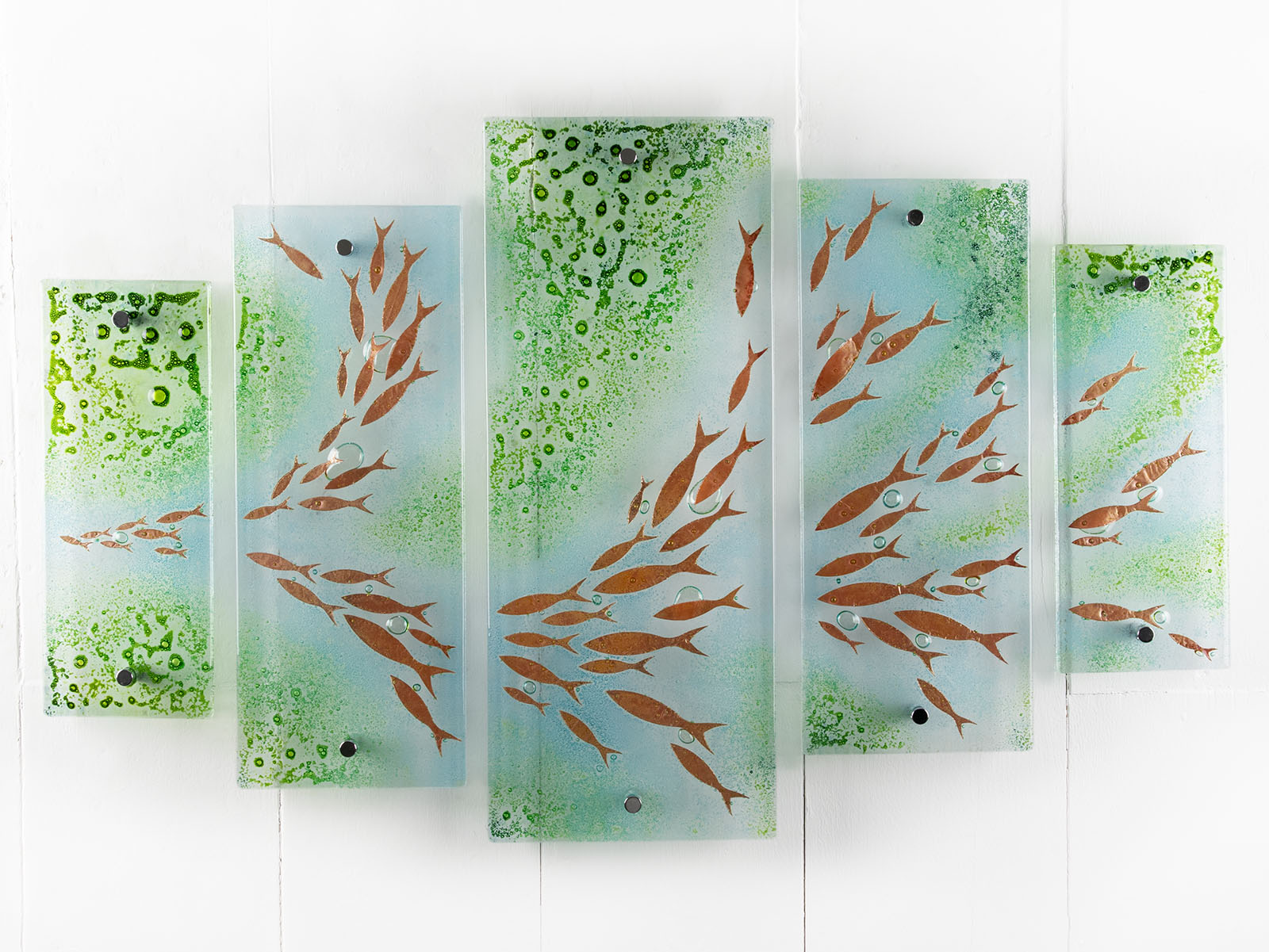 A fused glass quintych featuring a soft coastal colour-way with darting copper fish and bubbled detailing. An ocean inspired piece of wall art. 