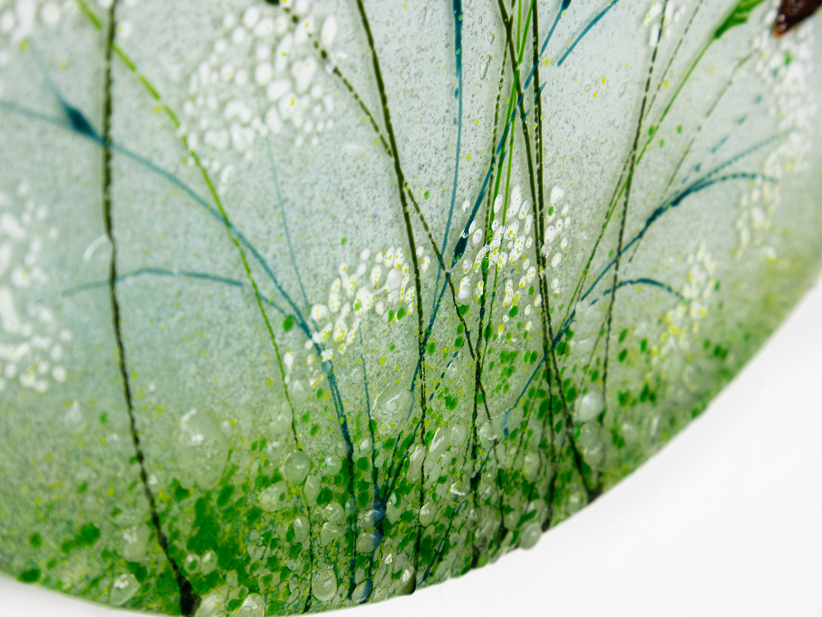 Close-up of a circular glass art piece featuring abstract green and white floral or grass-like patterns with clear bubble details on a white background.