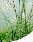 Close-up of a circular glass art piece featuring abstract green and white floral or grass-like patterns with clear bubble details on a white background.