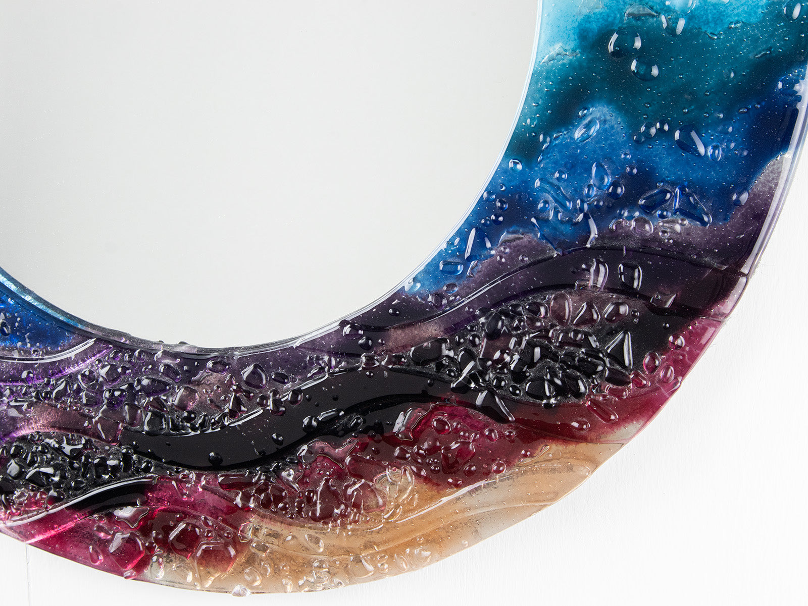 A detailed shot of a circular fused glass round mirror in a vibrant rainbow colour-way with soft waves and textured detailing. 