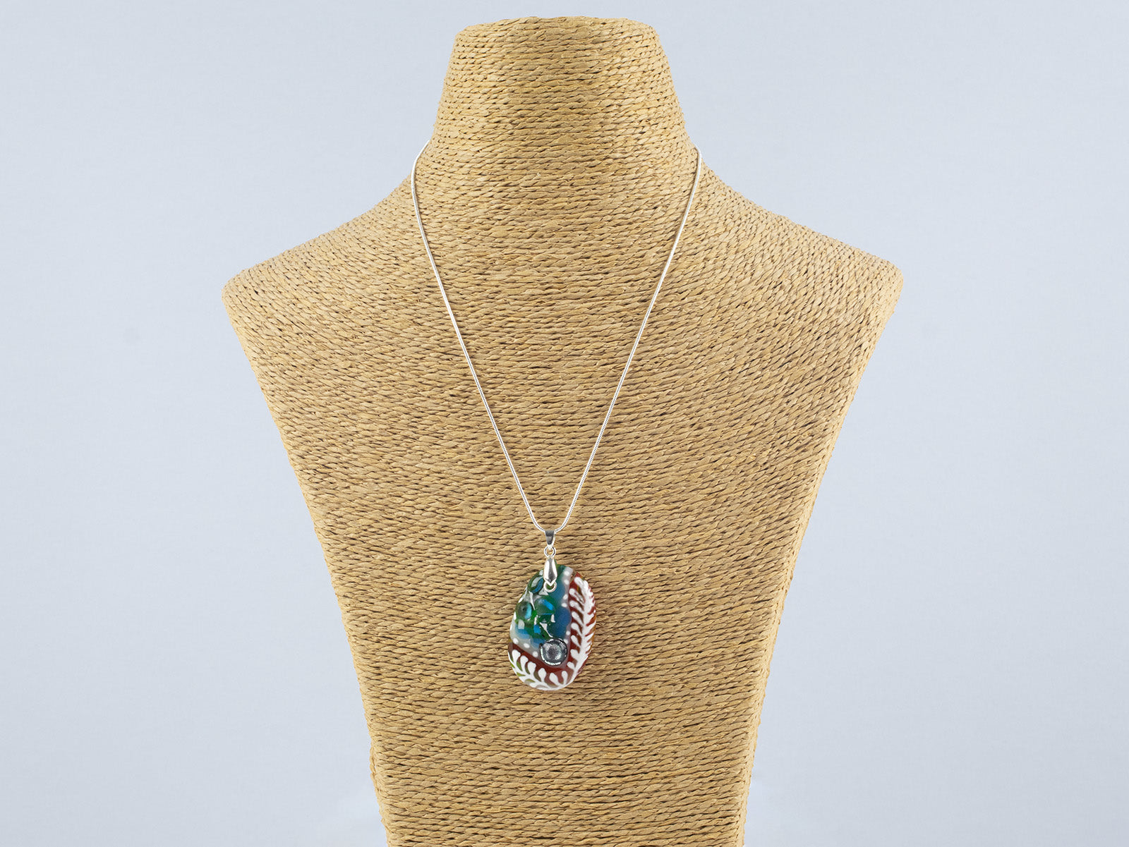 A fused glass pendant necklace on a sterling silver chain, handmade with soft coastal colours and a hint of red, with a hand-painted botanical design and a dichroic glass pebble. 