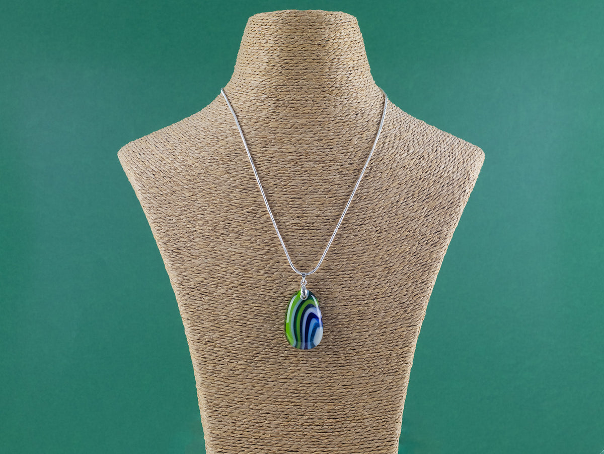 An artisan fused glass pendant necklace in coastal blues and greens, handcrafted in our studio. Finished with a sterling silver chain and fittings, a striking piece of wearable art. Shown on a mannequin. 