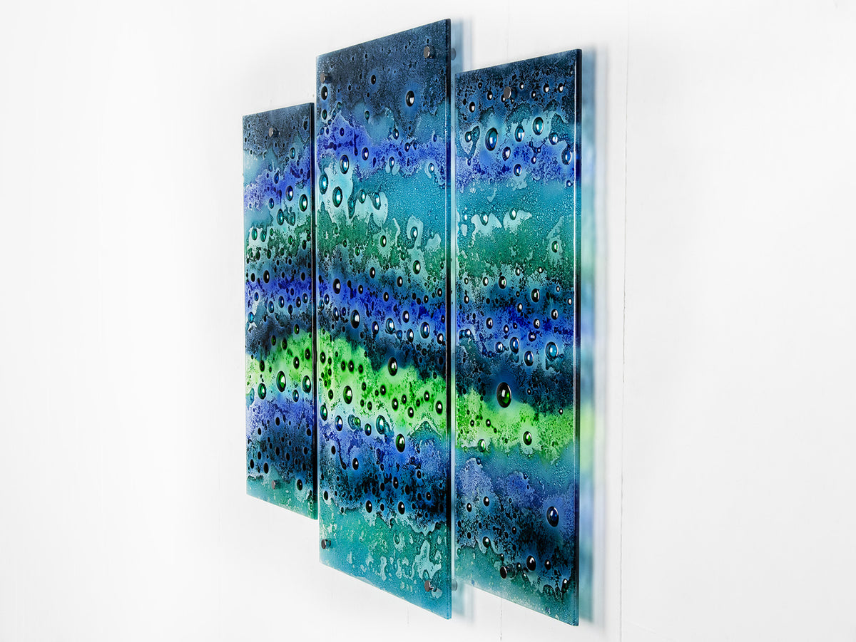 A fused glass staggered triptych featuring an ocean inspired design, with a blend of coastal blues and sea greens, handmade with organic bubbled texture and reflections.