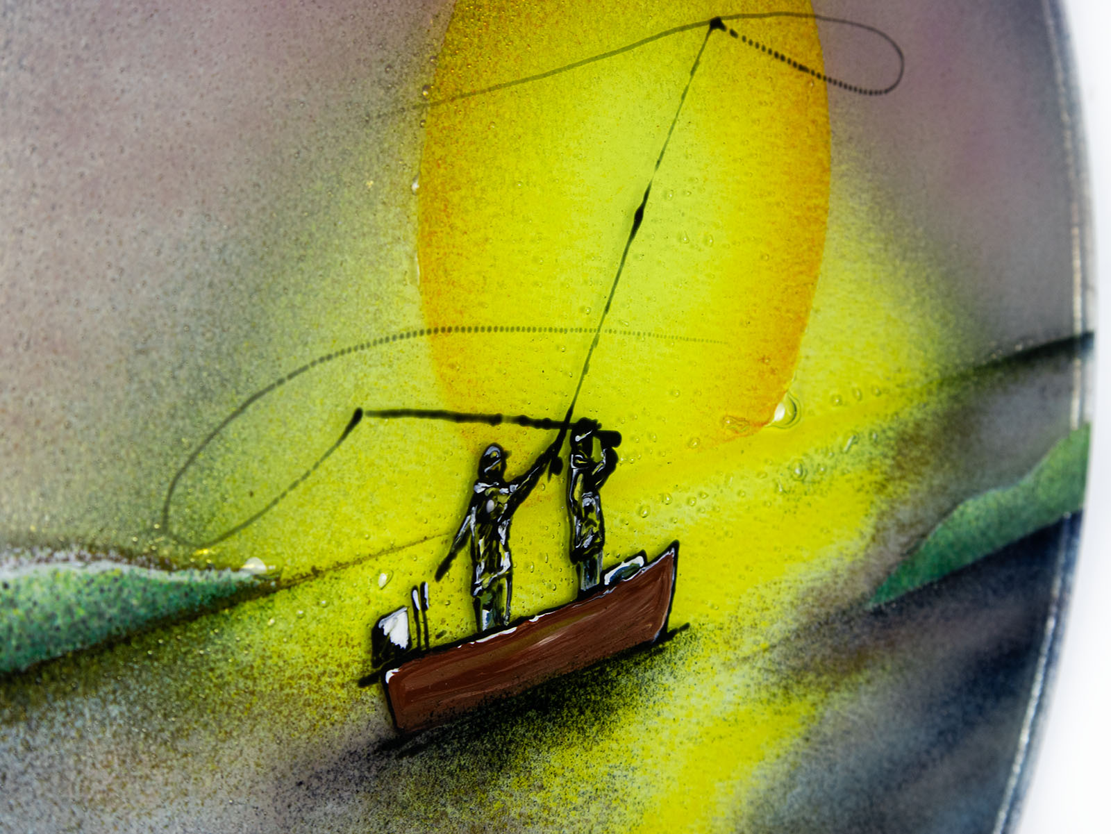 A close up of the painted details on a fused glass circular wall art panel featuring a design of a fisherman in a small boat casting a line in front of a large glowing yellow sun over a calm lake in natural, organic colours. 