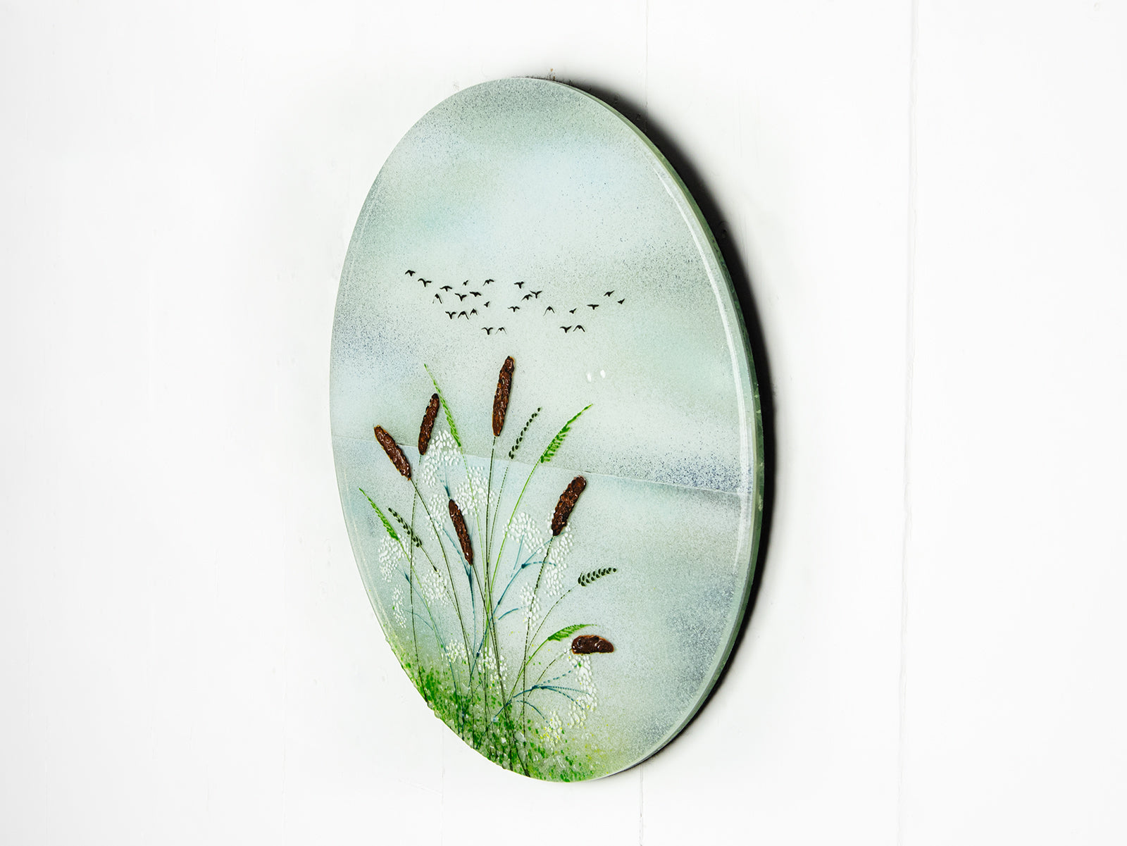 A circular fused glass wall art piece featuring a painted landscape with green reeds, brown seed heads, a pale blue sky, and small black birds, shown against a plain white background.