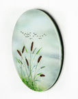 A circular fused glass wall art piece featuring a painted landscape with green reeds, brown seed heads, a pale blue sky, and small black birds, shown against a plain white background.
