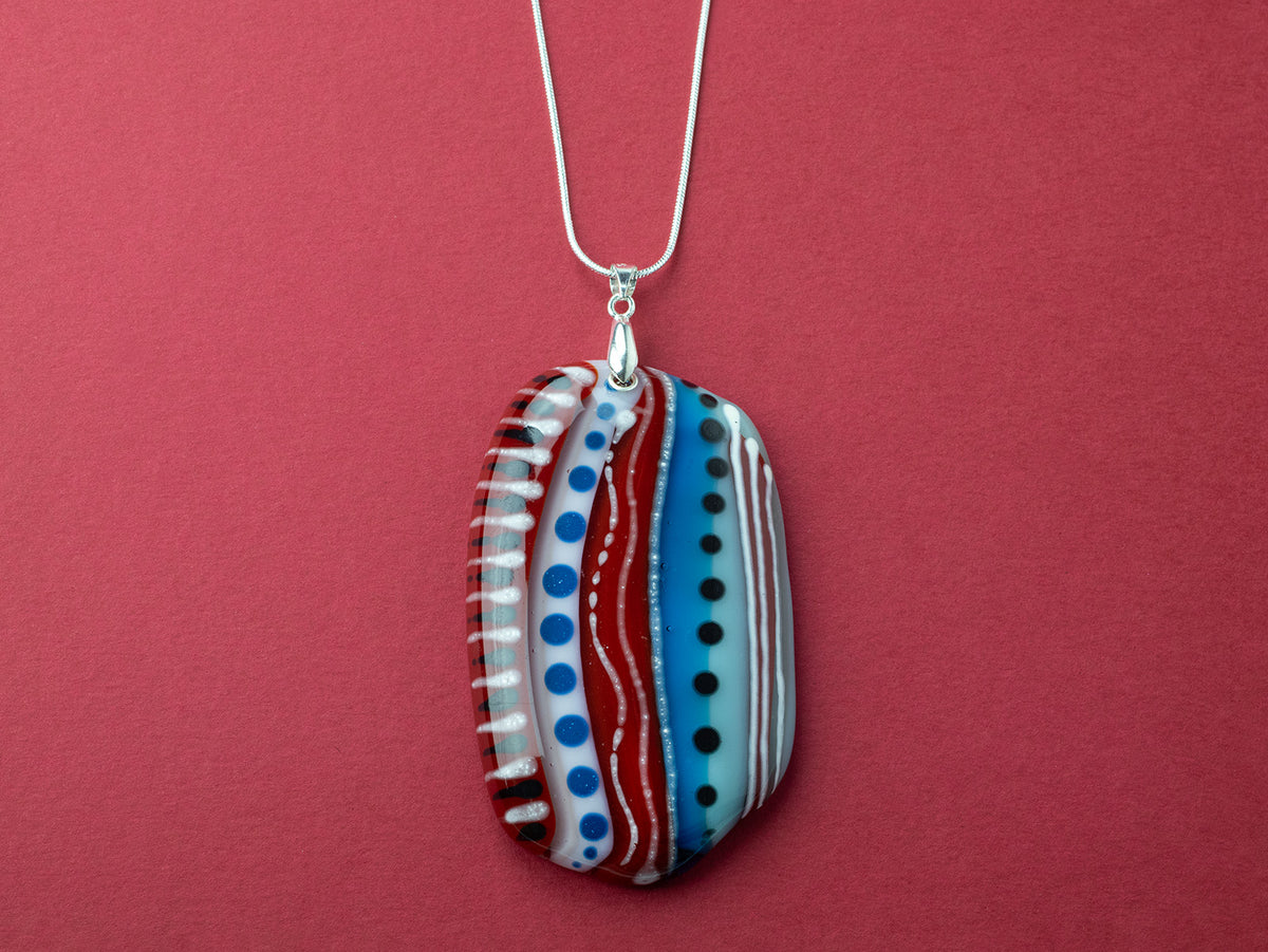 An extra large fused glass pendant necklace, handmade in rosey red and coastal blue hues, with hand-painted patterns and hung on a sterling silver chain 