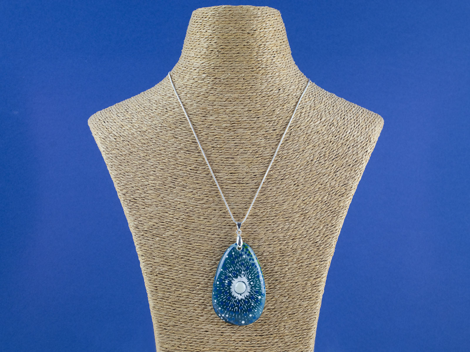 A teardrop-shaped fused glass pendant necklace featuring a blue and green sunburst pattern with white speckles and a central pearl inspired disc, suspended from a sterling silver chain.