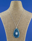 A teardrop-shaped fused glass pendant necklace featuring a blue and green sunburst pattern with white speckles and a central pearl inspired disc, suspended from a sterling silver chain.
