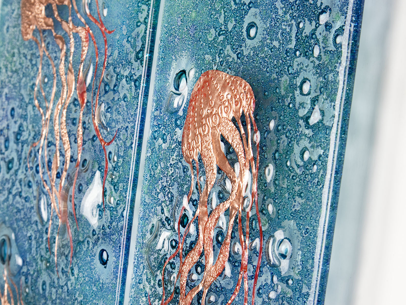 Close up of fused glass staggered triptych, handmade using copper, which has been hand etched into an intricate Jellyfish design, with bubbled marine blue background. 