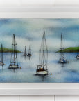 A hand-painted fused glass design of several sailboats anchored, designed in soft coastal colours and presented in a white frame.