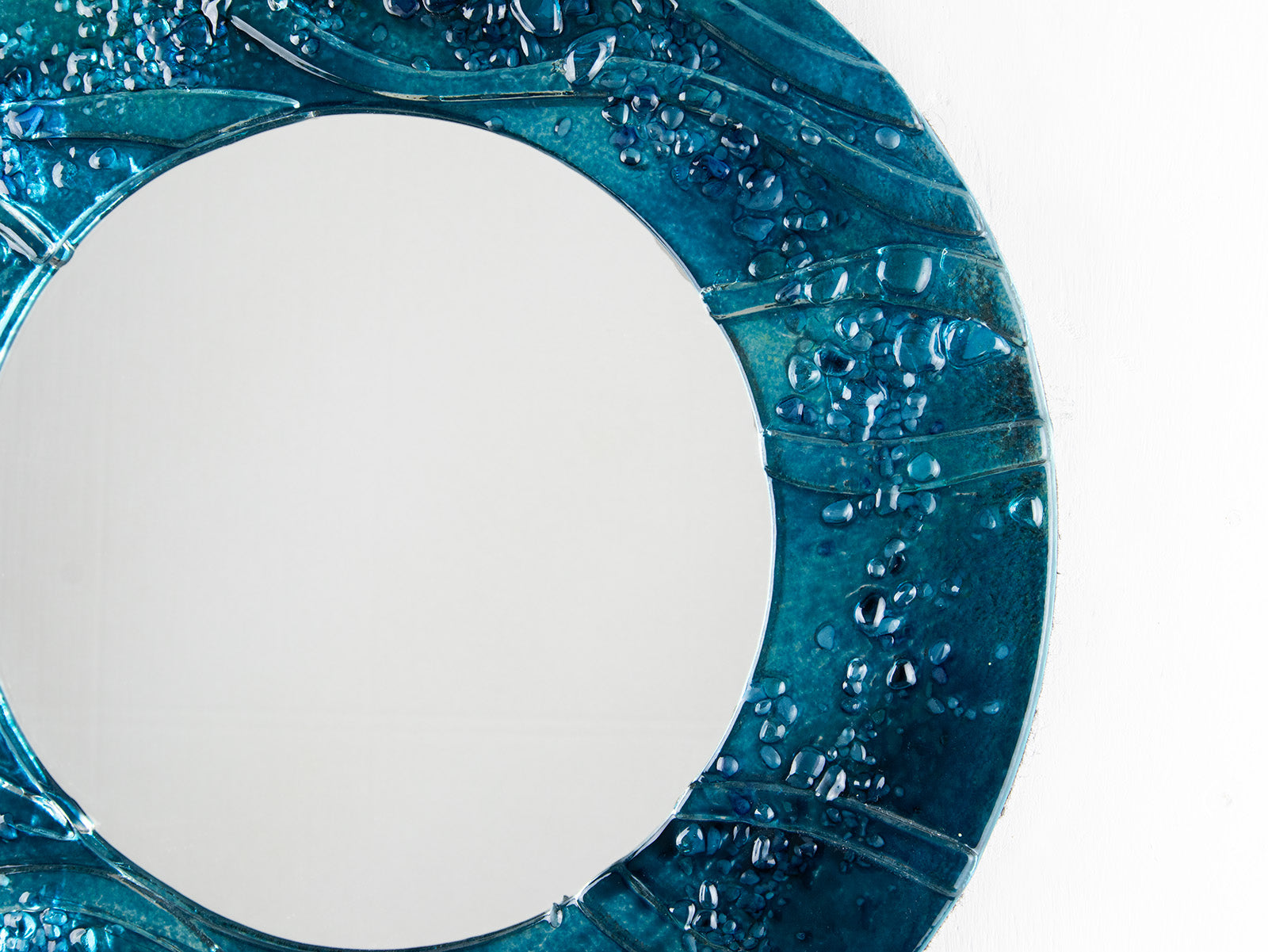 Close up of circular fused glass mirror, featuring teal blue textured waves, creating a coastal and ocean inspired wall art design. 