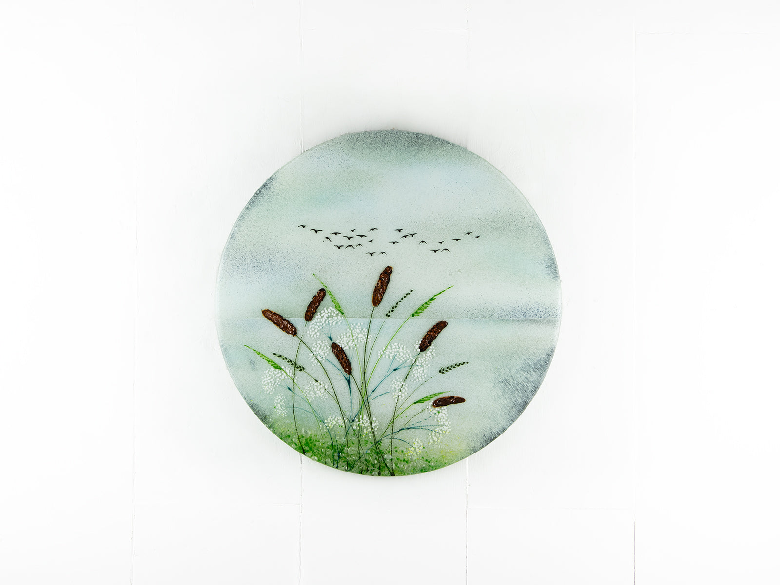 A circular fused glass wall art piece featuring a painted landscape with green reeds, brown seed heads, a pale blue sky, and small black birds, shown against a plain white background.