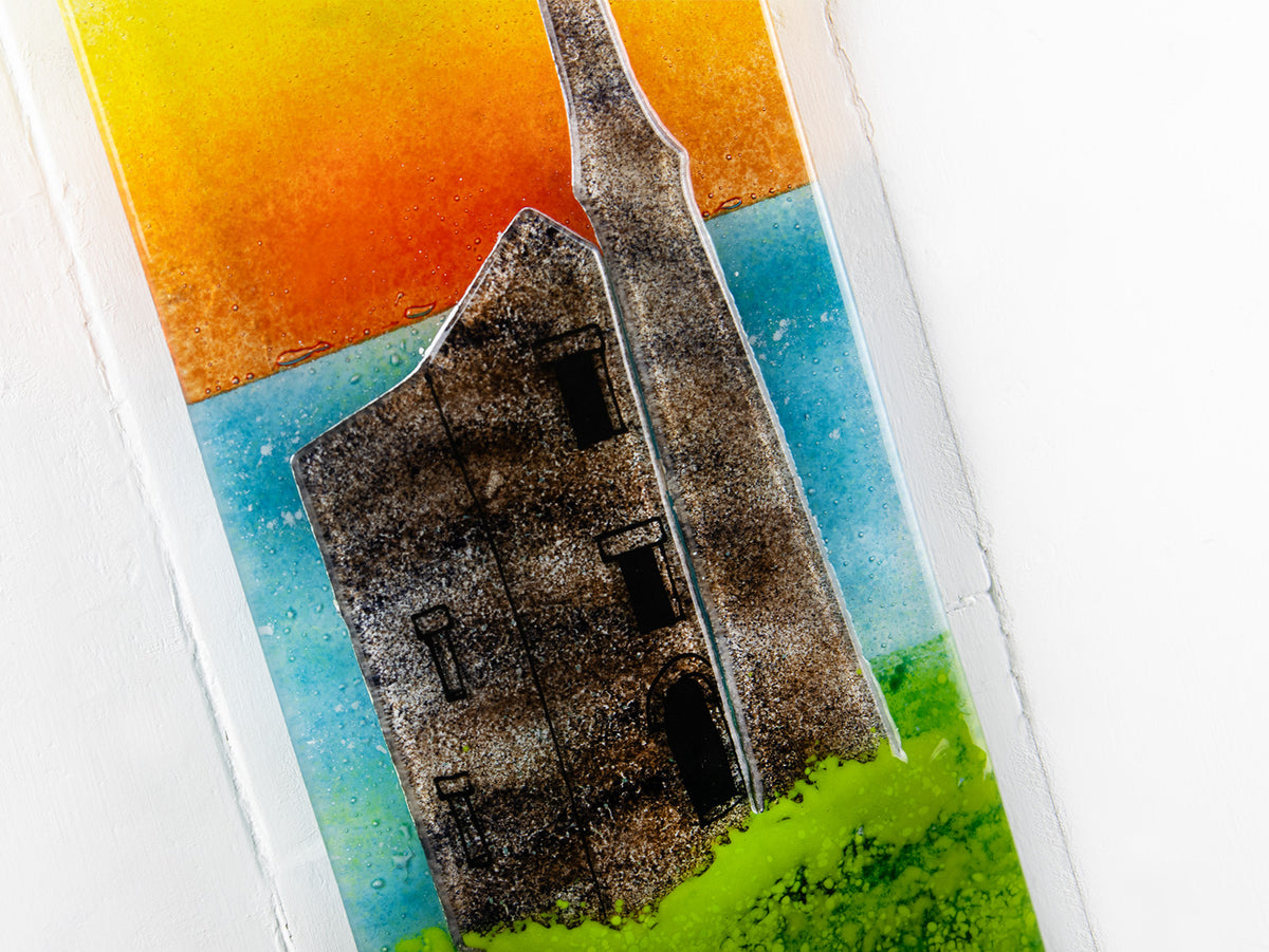 A close up of a fused glass wall panel, featuring a tin mine design, set against a vibrant sunrise in coastal colours. 