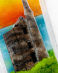 A close up of a fused glass wall panel, featuring a tin mine design, set against a vibrant sunrise in coastal colours. 