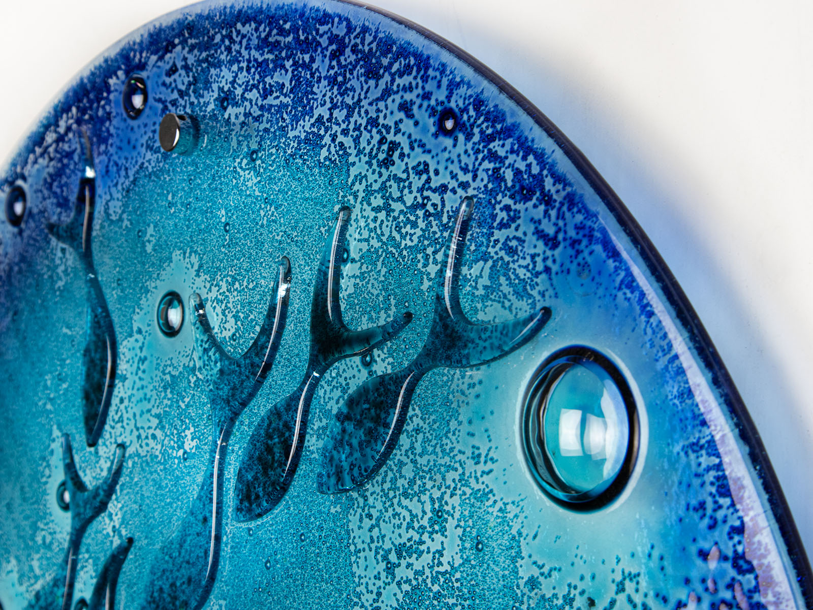A close up of a round fused glass wall panel featuring hand-cut glass fish and bubbles, designed in a marine blue colour-way.