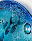 A close up of a round fused glass wall panel featuring hand-cut glass fish and bubbles, designed in a marine blue colour-way.