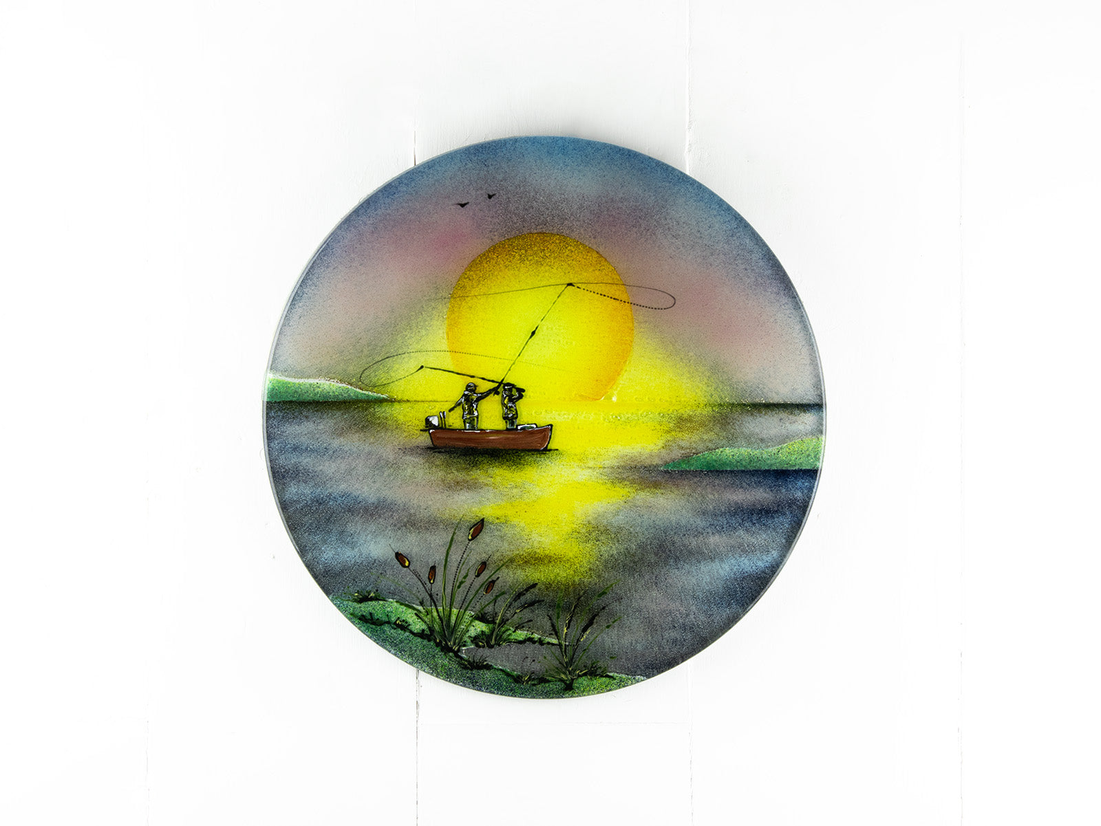 A fused glass circular wall art panel featuring a hand-painted design of a fisherman in a small boat casting a line in front of a large glowing yellow sun over a calm lake in natural, organic colours. 