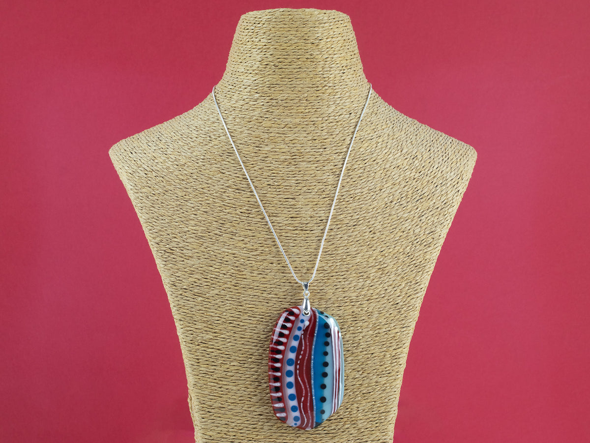 An extra large fused glass pendant necklace, handmade in rosey red and coastal blue hues, with hand-painted patterns and hung on a sterling silver chain 