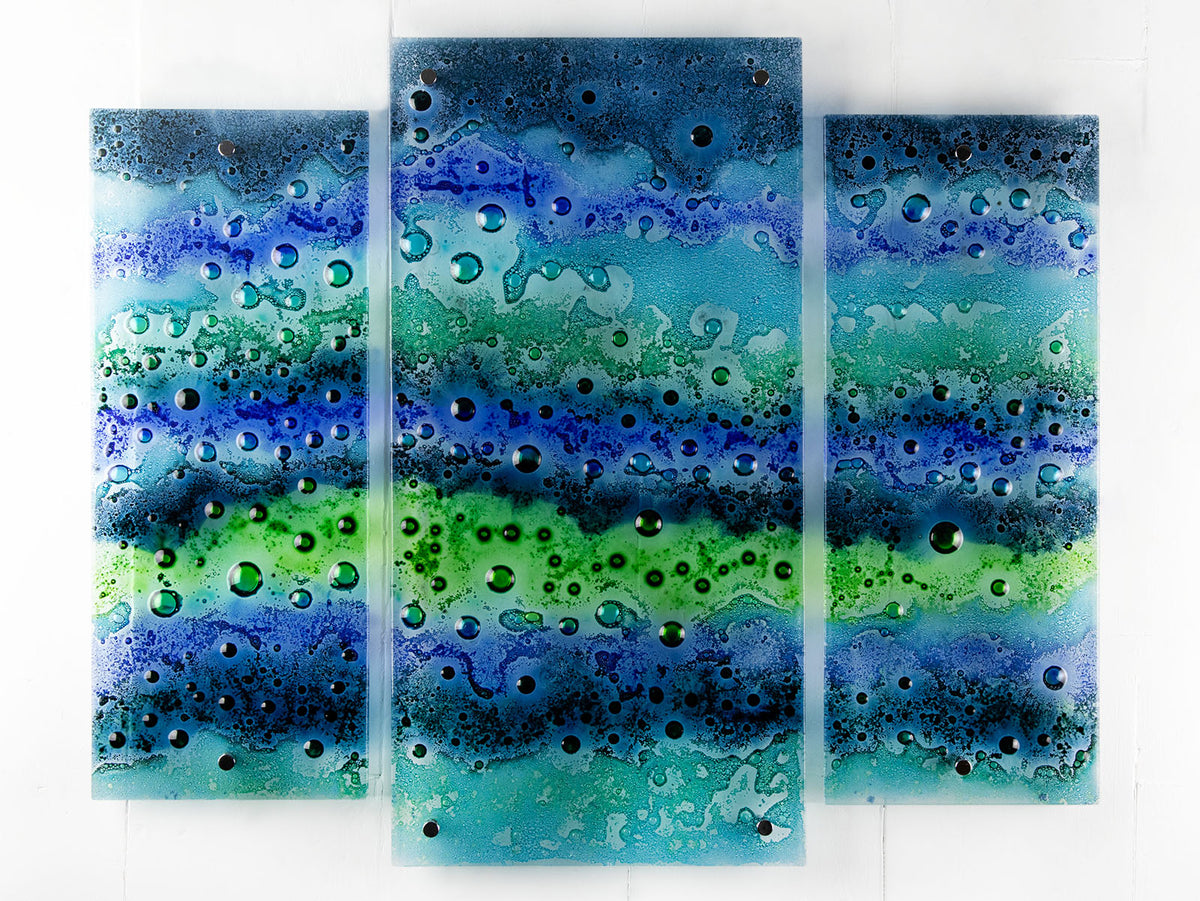 A fused glass staggered triptych featuring an ocean inspired design, with a blend of coastal blues and sea greens, handmade with organic bubbled texture. 