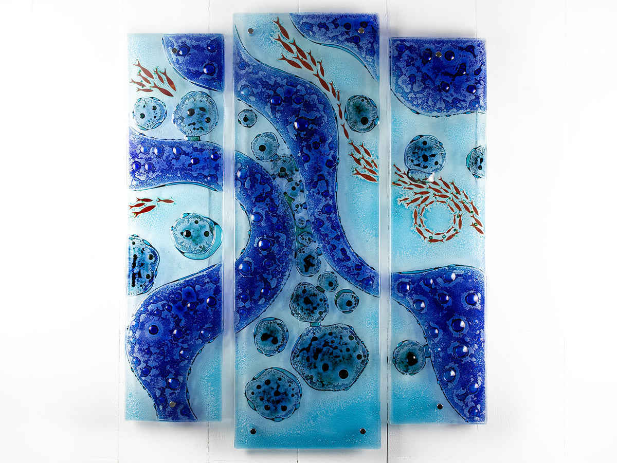 A fused glass staggered triptych wall display, handmade in bright ocean blues and aquas, in a coastal rock-pool design, featuring copper fish. 
