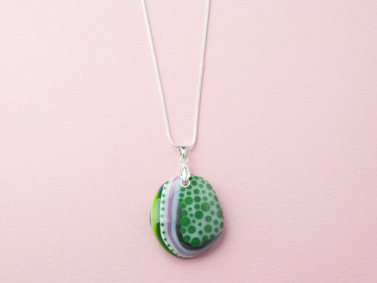 A fused glass pendant necklace, handmade in sea greens and blushed pinks, with hand-painted patterns, hung on a sterling silver chain. 