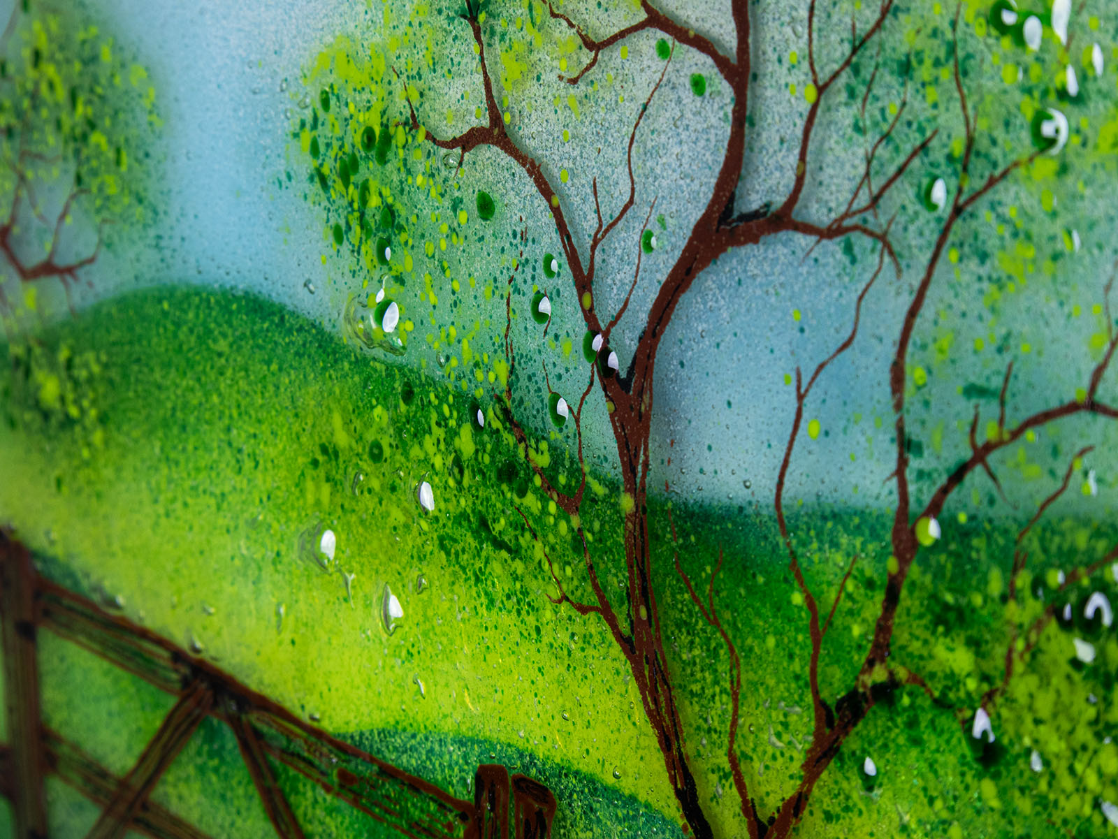 Gorgeous detailing featured on a circular fused glass wall art piece featuring a painted landscape with green trees, a wooden fence, rolling hills, and a dirt path under a light blue sky.