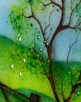 Gorgeous detailing featured on a circular fused glass wall art piece featuring a painted landscape with green trees, a wooden fence, rolling hills, and a dirt path under a light blue sky.