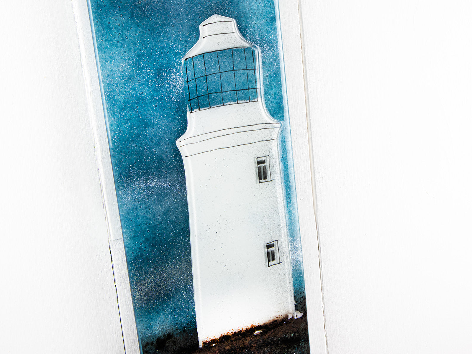 Close up of fused glass wall panel featuring a Cornish lighthouse design with a teal blue background and full moon design. 