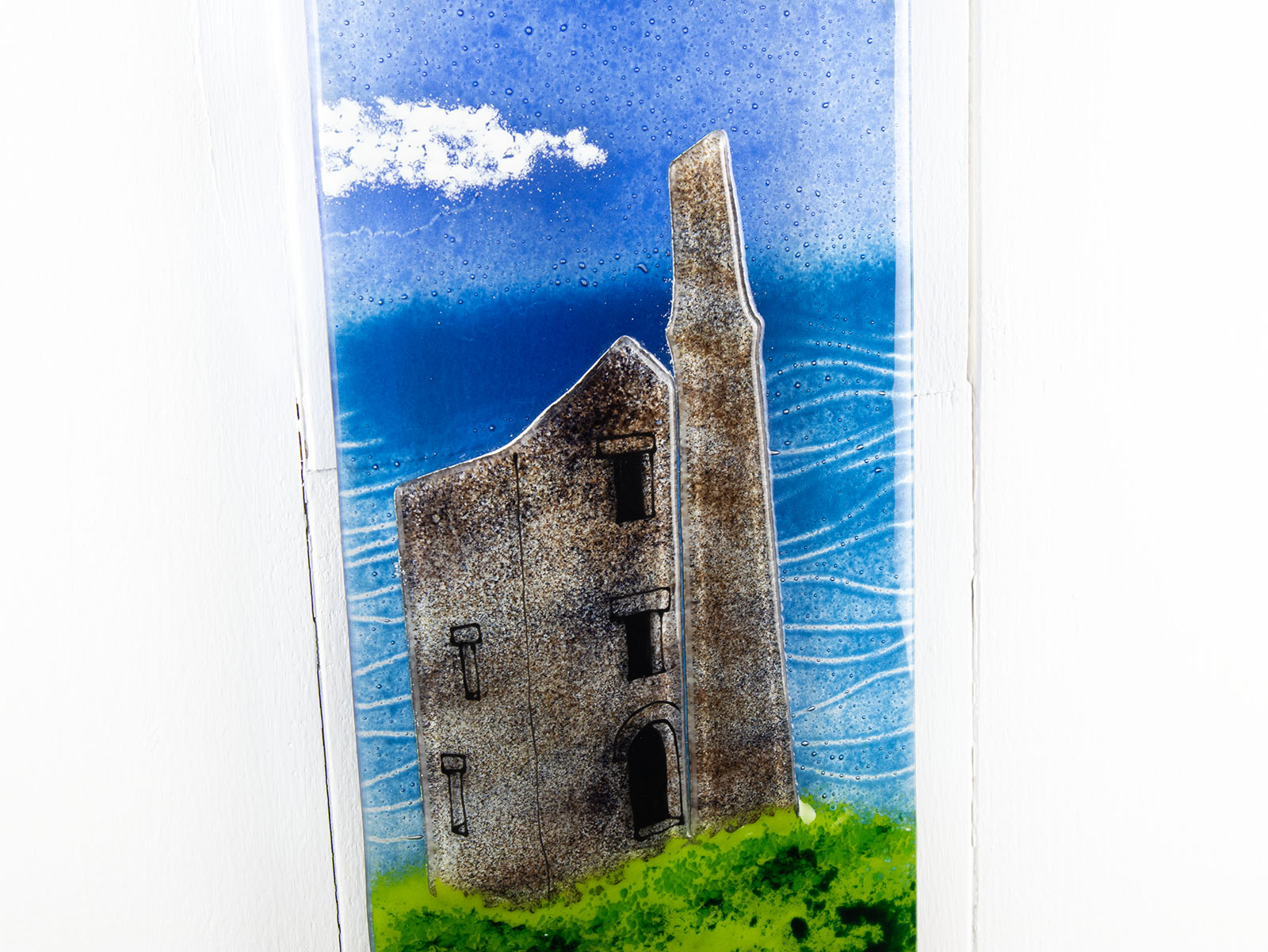 Close up of a fused glass wall panel featuring a Cornish lighthouse sat against a coastal blue design.  