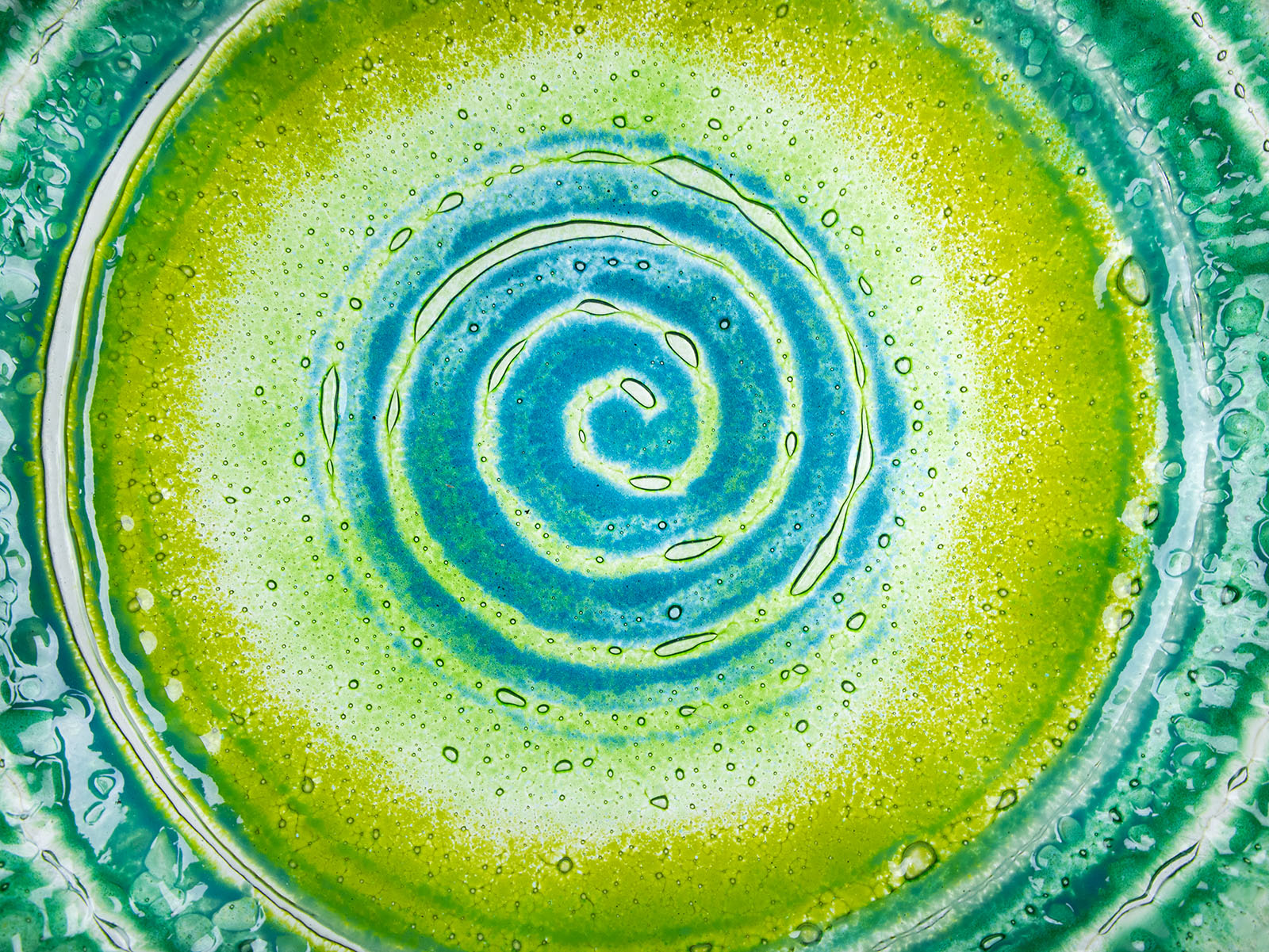 Close up of fused glass round dish featuring bright sea greens and swirling aqua detailing. 