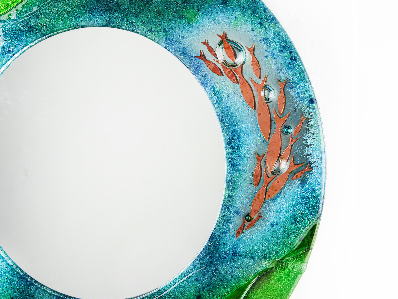 Close up of fused glass round mirror, featuring coastal design in sea greens and blues with copper fish. 