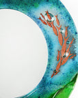 Close up of fused glass round mirror, featuring coastal design in sea greens and blues with copper fish. 