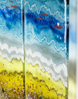 Close up of fused glass triptych depicting a beach scene with blue water, yellow sand, and red, copper fish.