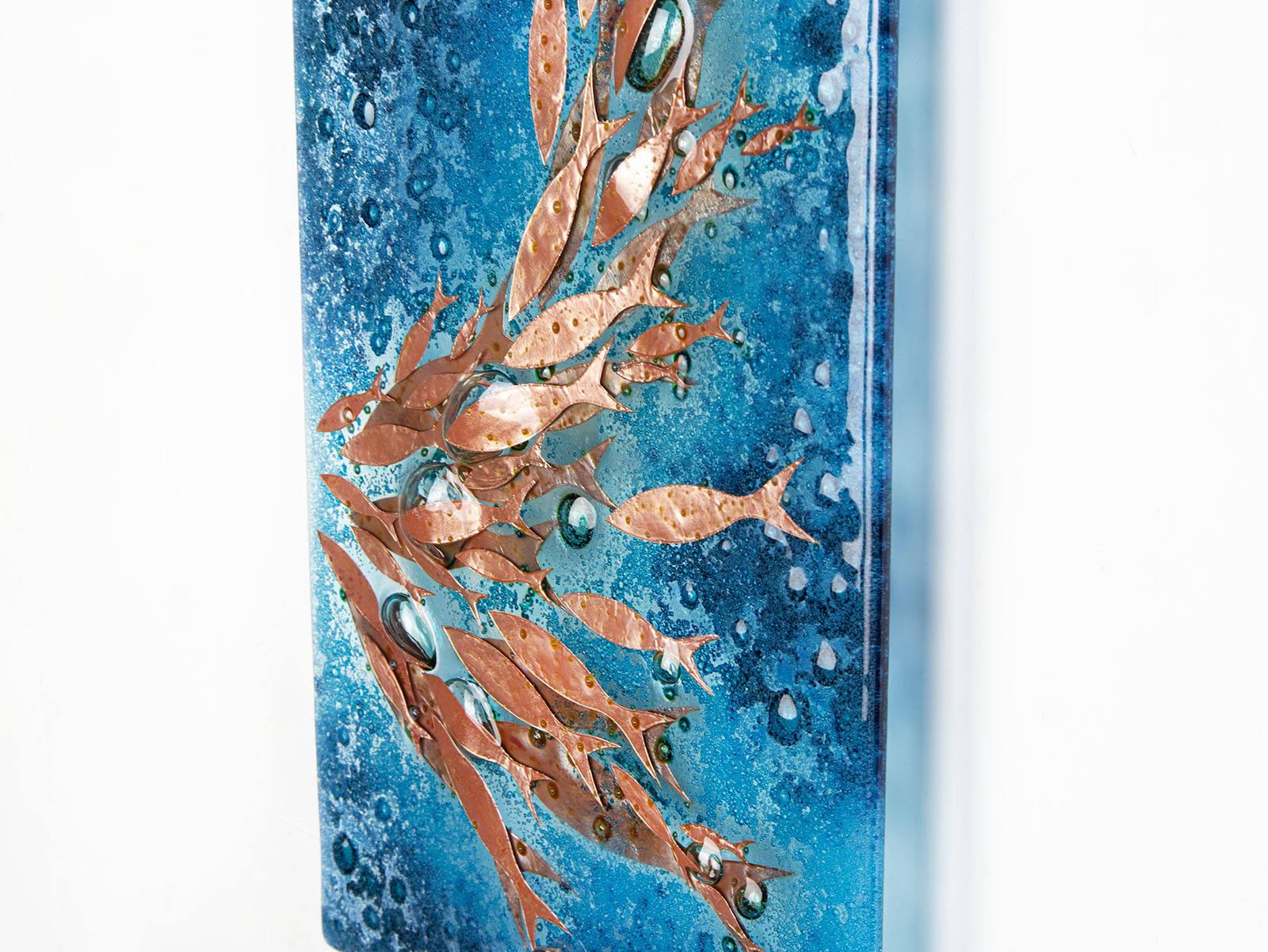 A close up of fused glass wall panel with bubbled blue colours and swimming copper fish. An organic and ocean inspired piece of wall art. 