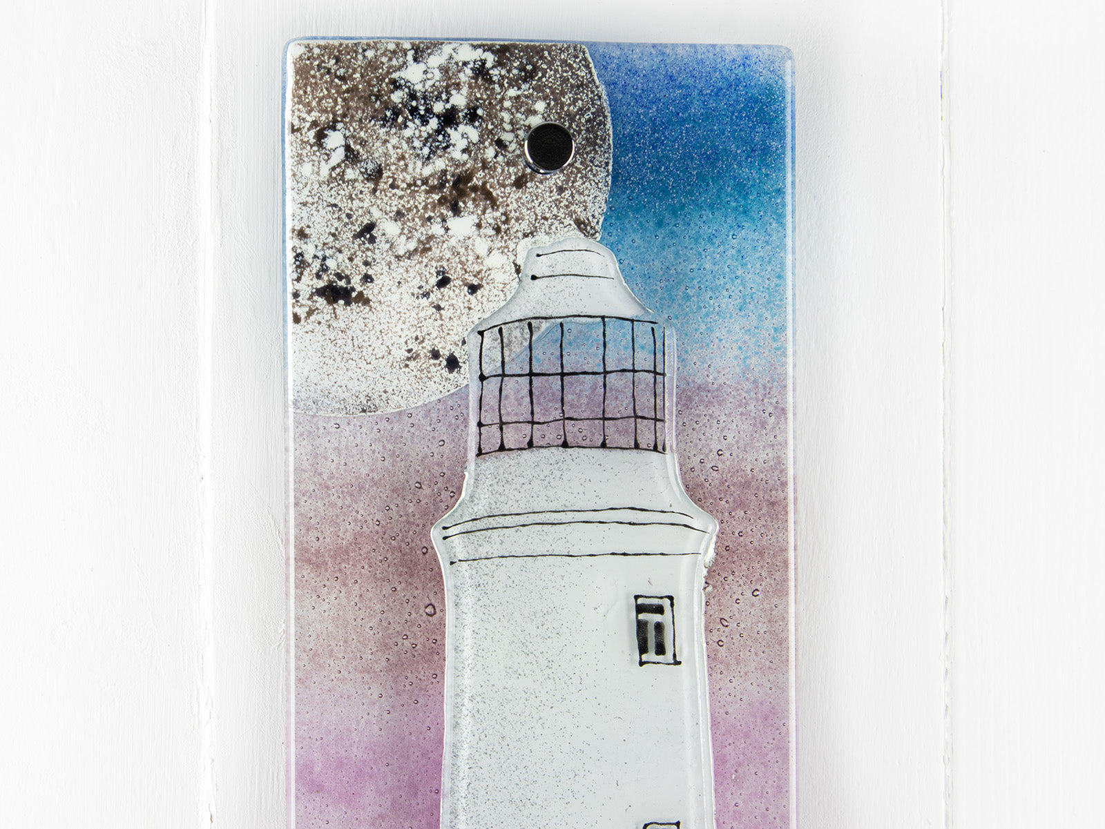 Details of small fused glass wall panel with a Cornish lighthouse and full moon design, with coastal hues and a blushed pink sky. 