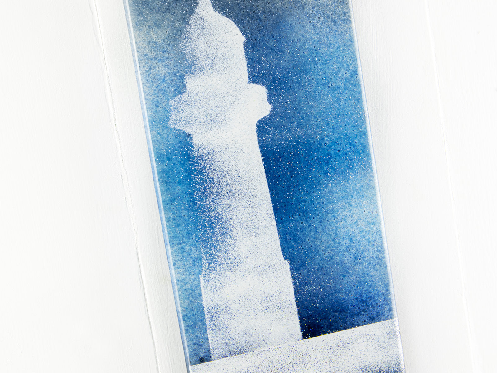Closeup of a fused glass wall panel with a soft white lighthouse design against a teal blue background. 