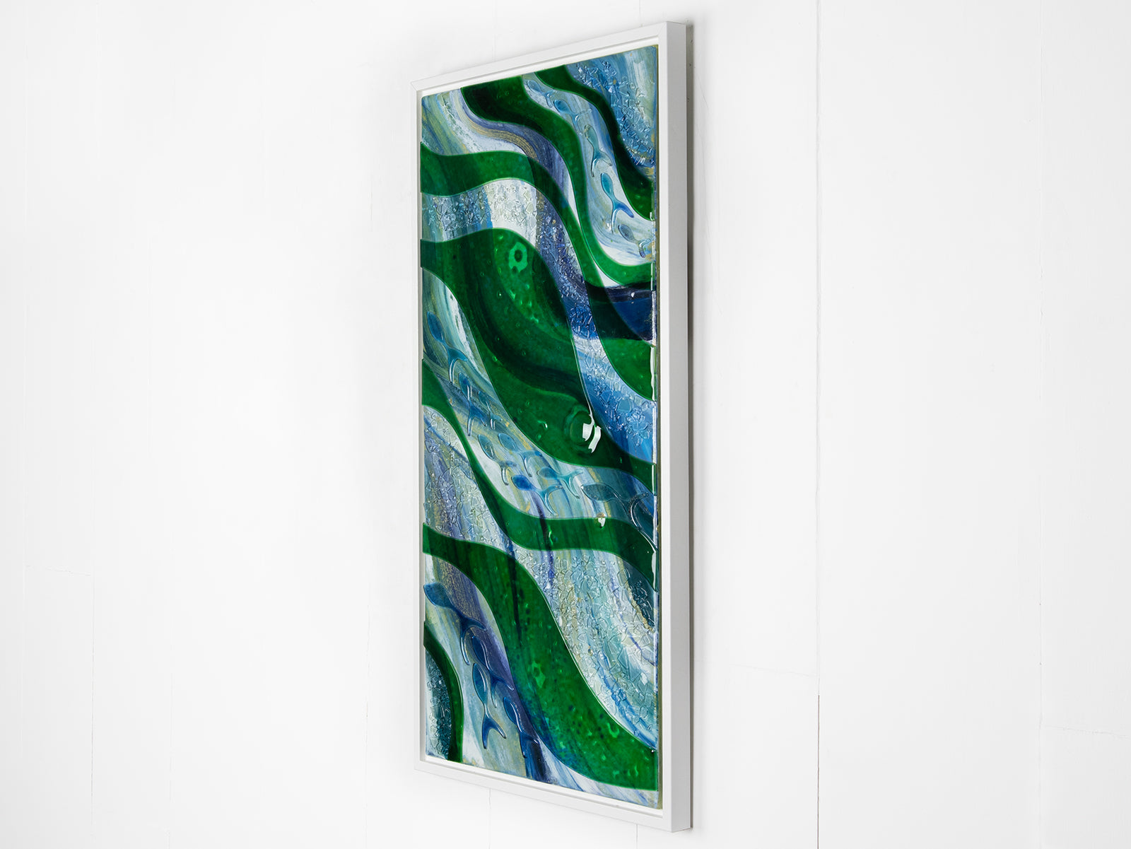 Side on image of a fused glass oblong art frame with coastal greens and textural blues. Hand-made with lass fish layered on and a white frame.