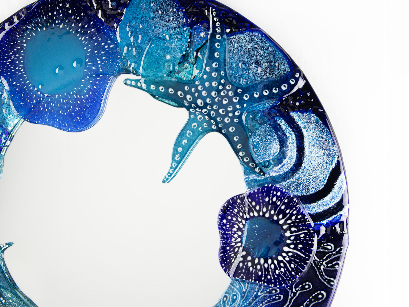 A close up of round mirror, made from fused glass with hand-painted ocean designs. Coastal blue colour-way, handmade and bespoke wall art.